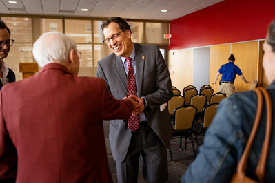 A man in a suit is smiling really big and giving another man an enthusiastic two handed hand shake. They might be friends. My guess is the man wants to be friends with this guy. 