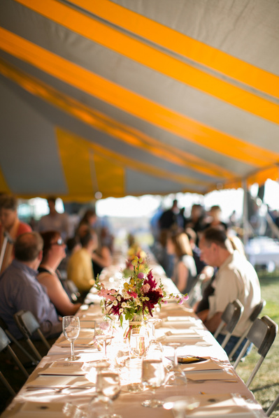 A long table of people sitting getting ready to eat at an outdoor meal. The tent they are under has yellow strips on the roof. 