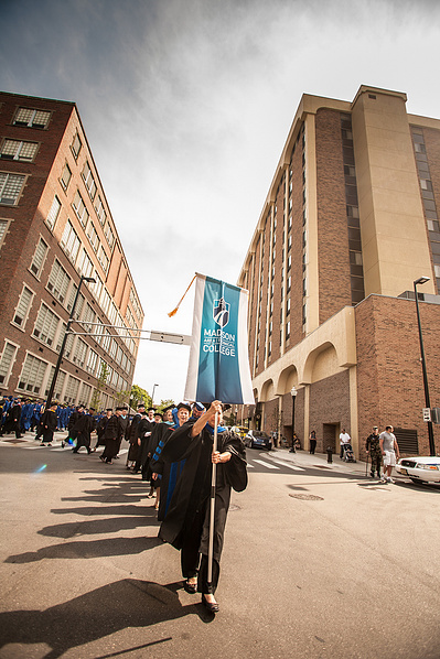 A group of Madison College students walk between 2 tall buildings, holding a banner on the way to their graduation.