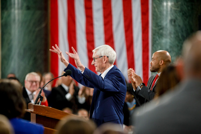 Tony Evers is talking to a crowd at the State Capitol in Madison WI. There is a flag behind him--so very patriotic. 