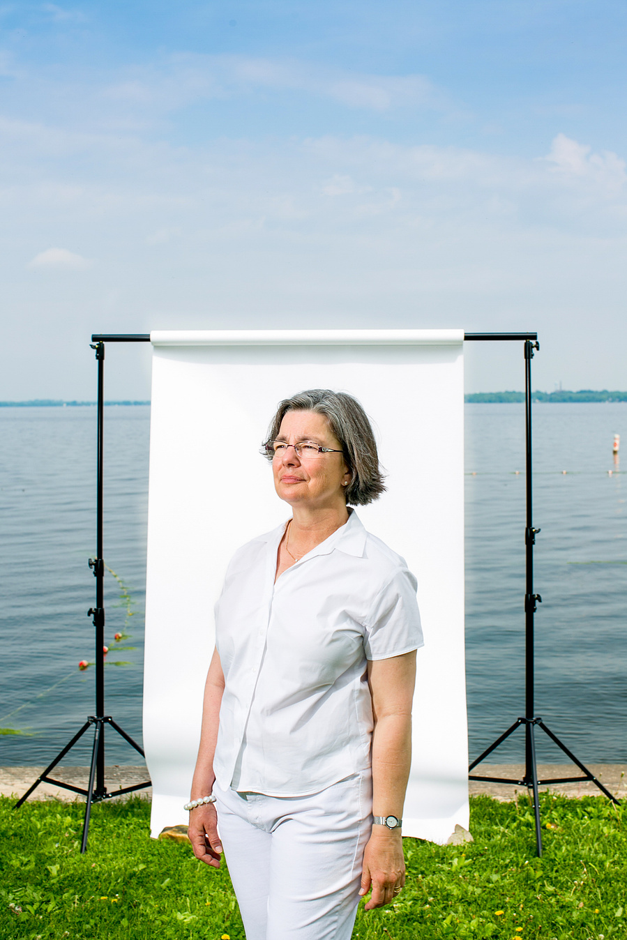 A gal wearing all white is posed with a  background stand in background with the lake behind the background stand. 