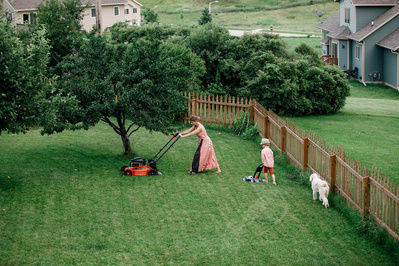 A fun photo of a mother mowing her lawn while her children follow along behind her. Family documentary photo.