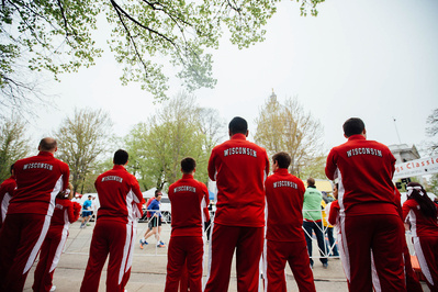 A group of students from the UW Madison are wearing red track suits with the word "Wisconsin" on the backs on the suits" They are watching a marathon. 
