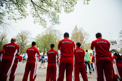 A group of students from the UW Madison are wearing red track suits with the word "Wisconsin" on the backs on the suits" They are watching a marathon. 