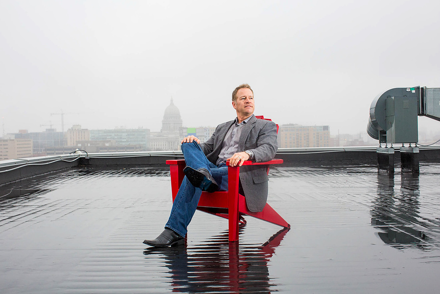 A businessman is sitting on a red chair on the top of a building in downtown Madison. The Capitol is in the background. 