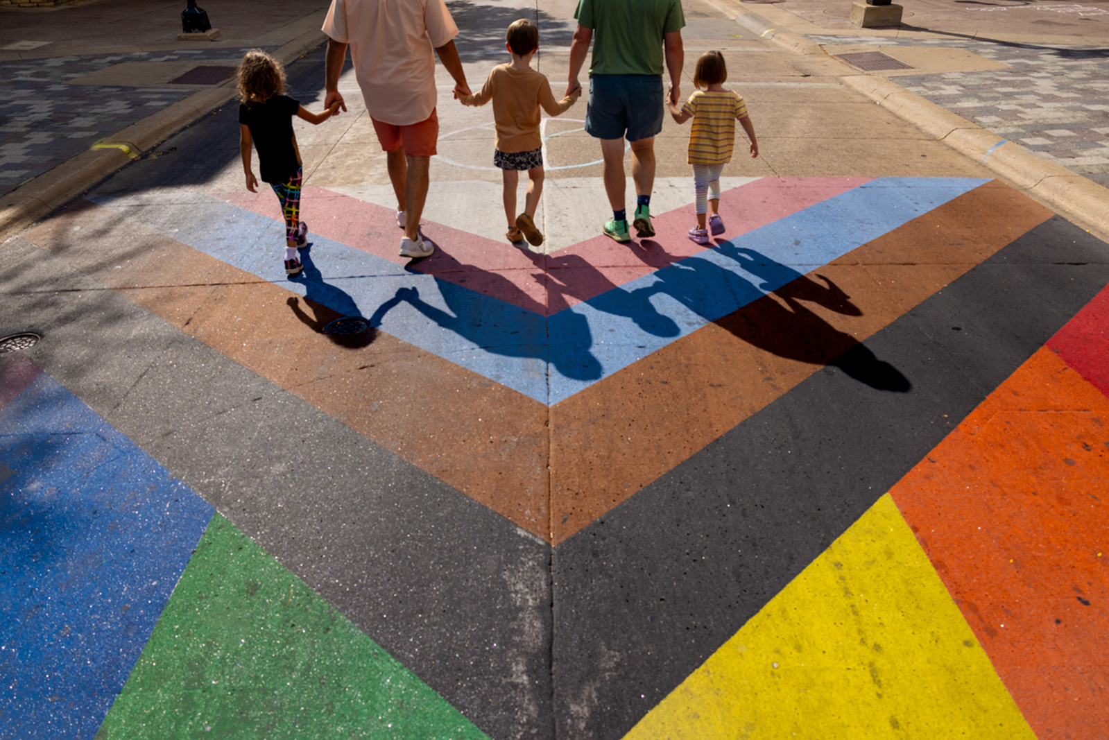 A documentary photograph of fathers and children walking along State Street near the capitol building in Madison, Wisconsin.  The Progress Pride Flag also known as the Inclusive Rainbow Flag frames the family enjoying a walk on the Capitol Square.