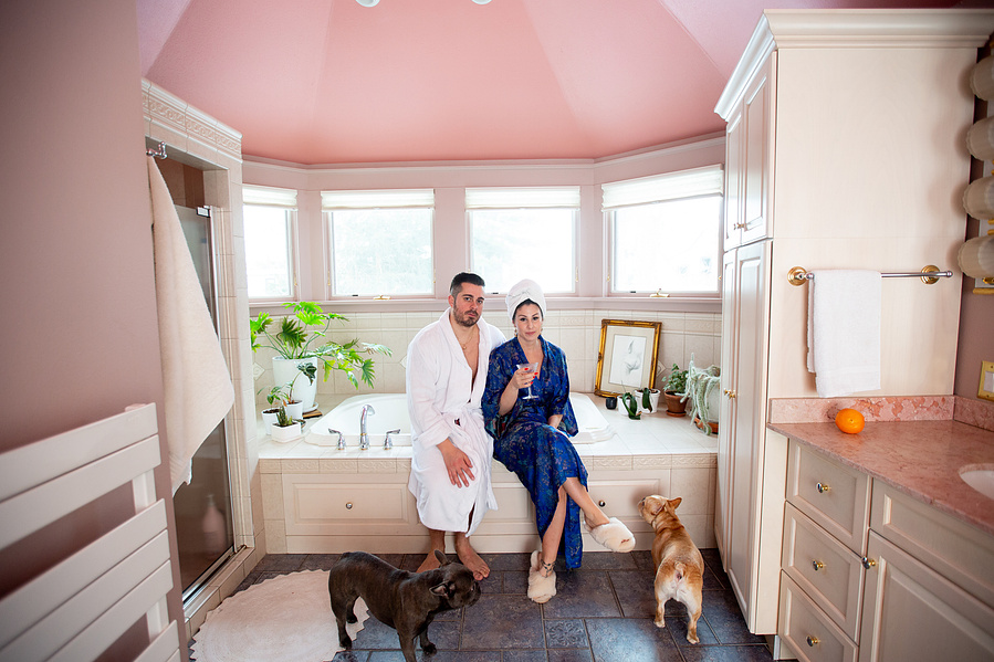 A luxurious couple pose in their pink bathroom wearing bathrobes with their french poodles.