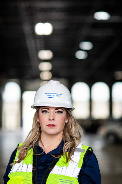 A women is posing for the camera wearing a hard hat and utility vest. 
