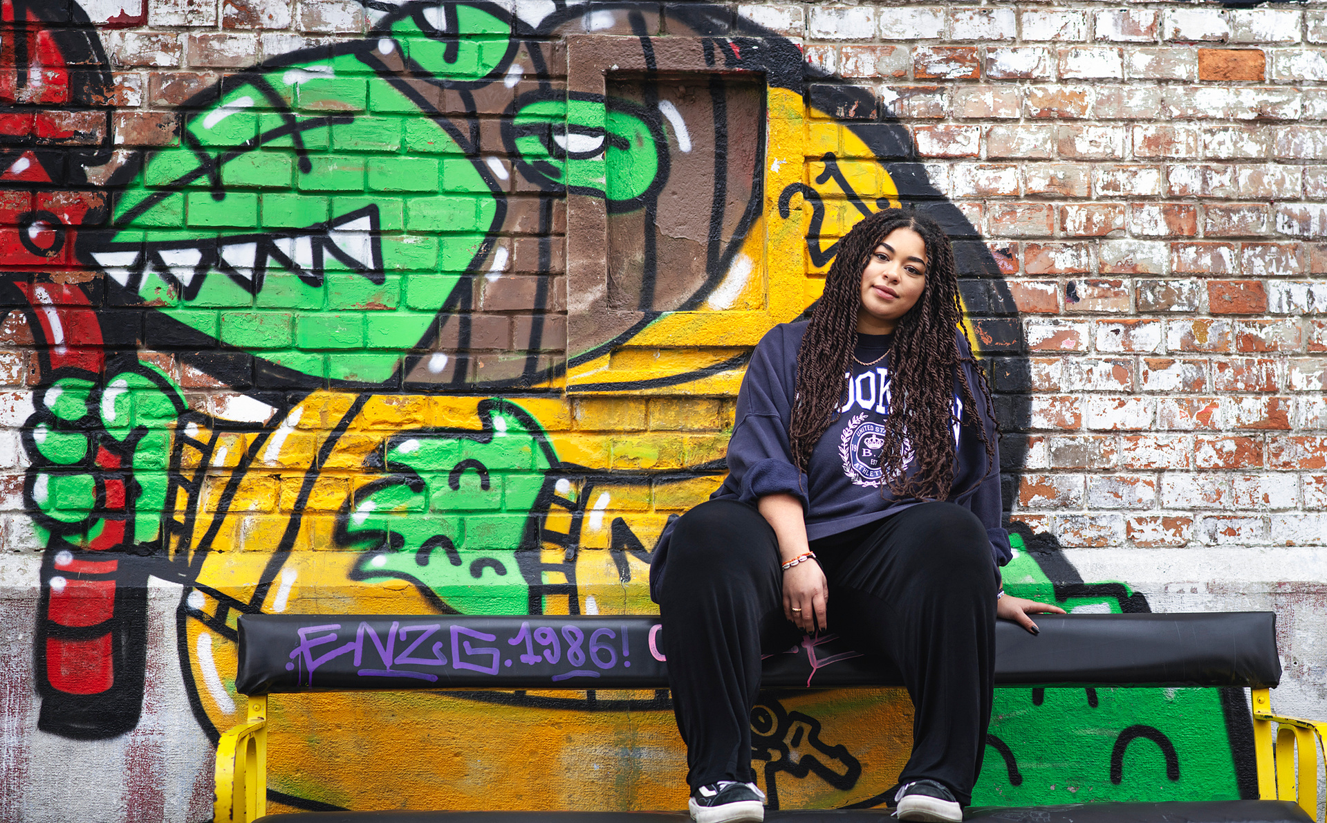 Junge Frau sitzt entspannt an einer bunten Graffitiwand – urbanes Portrait mit kreativem Street-Art-Hintergrund. Niederösterreich, Tulln, Krems an der Donau