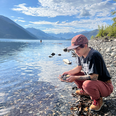 River Kenji is crouched along the rocky shore of a creek in Greenwood, BC. They are wearing a cap, black t-shirt with rust coloured pants and hiking boots. Behind them is a person on the horizon paddling away in a kayak with mountains in the background.