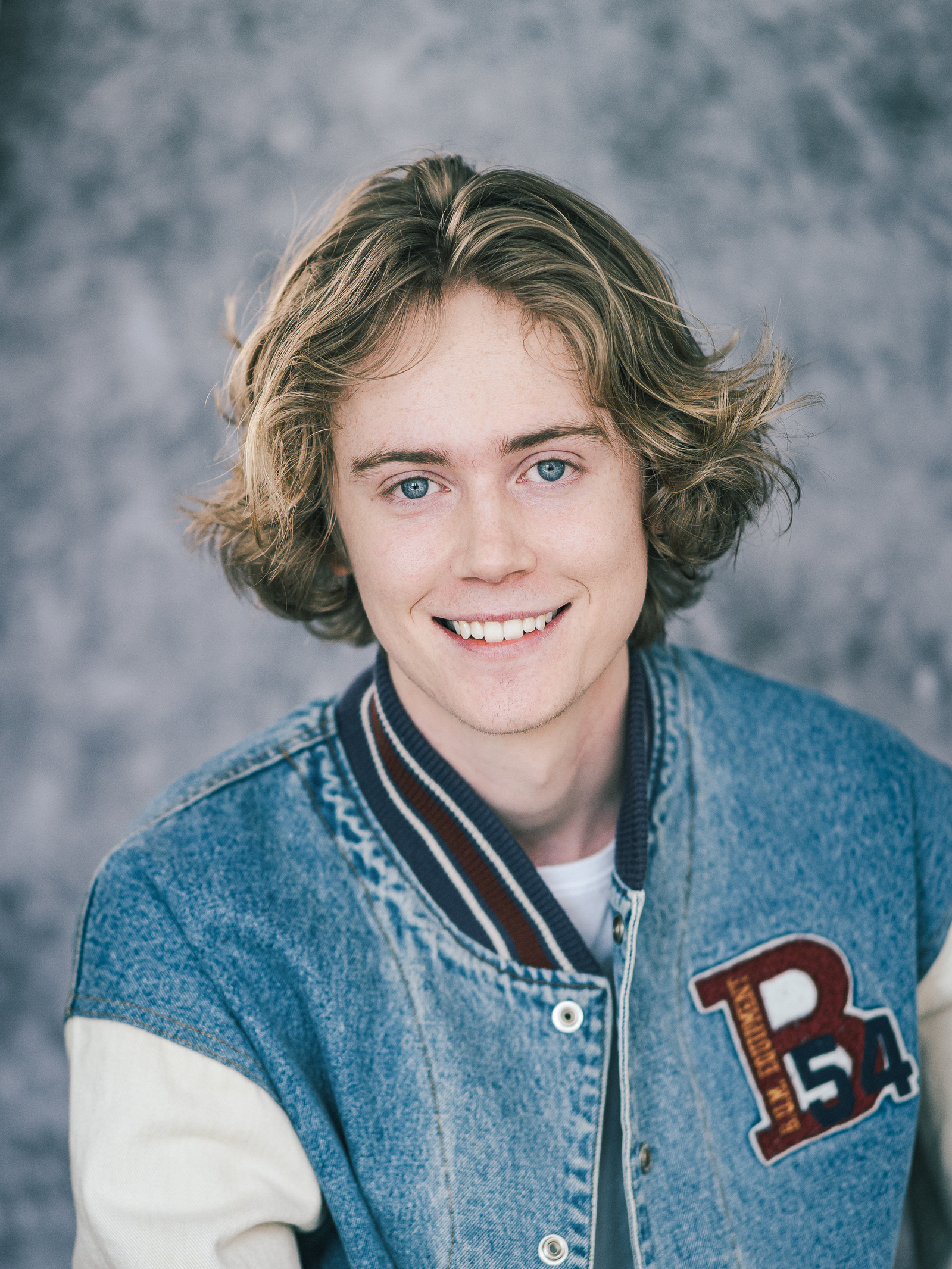 Headshot of a blonde male actor wearing a blue jacket against a grey background