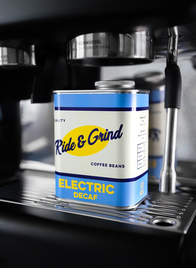 A  photograph of a coffee tin placed on a coffee espresso machine next to the steam wand. The tin has the brands logo on the front which reads "Ride & Grind" and the name of the coffee beans "Electric Decaf" Coffee roasted in Edinburgh Scotland.