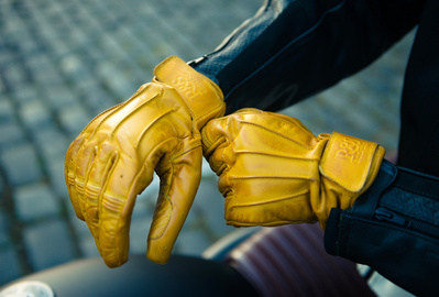 Motorcycle Gloves that are yellow worn by a rider sitting on a black motorbike parked on cobbled streets.