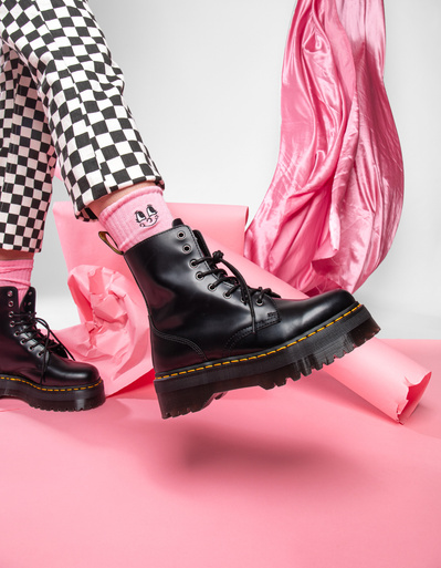 Dr Martens Black boots with yellow stitching photographed in a studio on a pink backdrop with pink designer socks and checkerboard trousers on a female model.