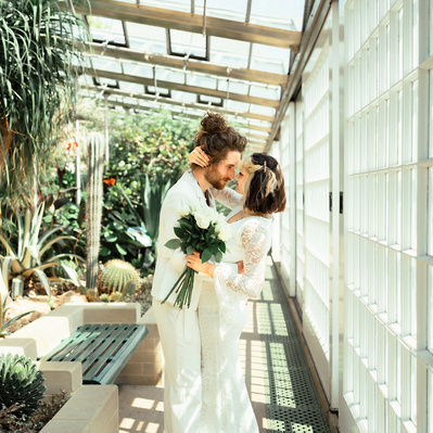 Alternative couple having wedding photos taken in a botanical gardens with plants in the background. She holds white roses and wears a white alternative bohemian dress.