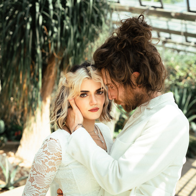 Alternative couple having wedding photos taken in a botanical gardens with plants in the background. She wears a white alternative boho dress and he has a white suit.