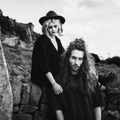 Newly engaged couple having an engagement photo shoot at the roaches in the peak district. Alternative couple wearing black with tattoos and piercings with her sat on a stone wall behind him.