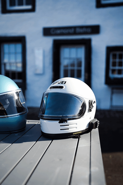 A Shoei motorcycle helmet next to a biltwell gringo helmet both on a bench outside a pub in scotland on a sunny day after a ride.