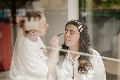 Are you colour photograph through a window reflection of a Bride having her make up applied on her wedding day?