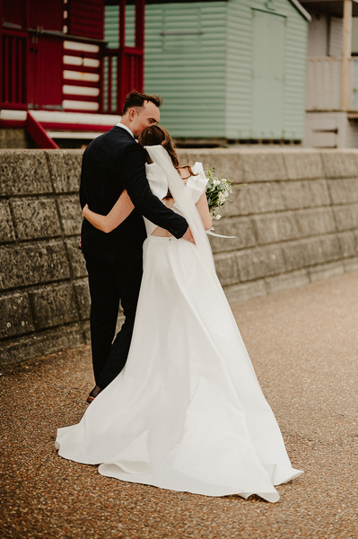 bride and groom walking along Whitstable promenade shot from behind hugging