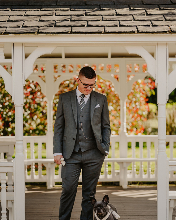 A natural, unposed photo of a groom standing outdoors, exuding a relaxed vibe, with his loyal dog casually resting at his ankle