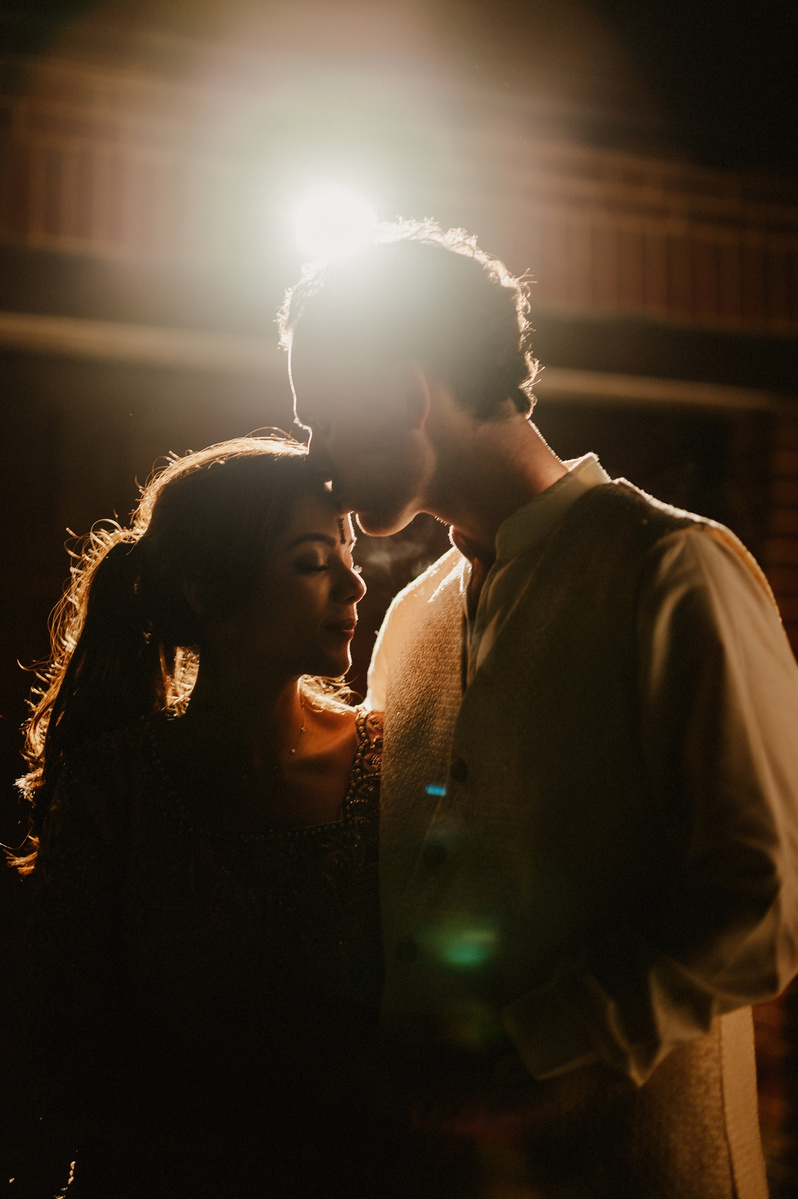 relaxed low-light wedding photography portrait at a shallow depth of field after a garden wedding reception in kent, the couple are only lit by a garage flood light creating a beautiful lens flare whilst the groom kisses the bride on the forehead
