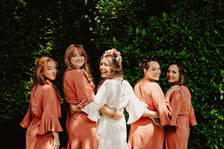 Bridal squad posing displaying their robes in front of a green hedge on wedding morning