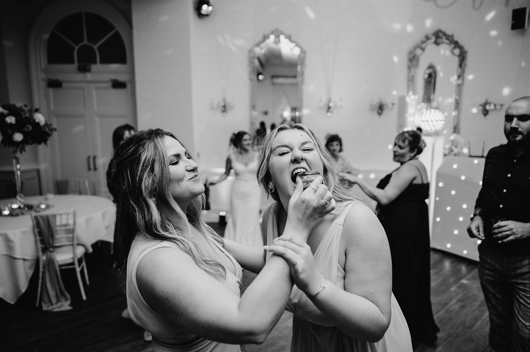 A candid black and white photo of two bridesmaids on the dance floor, one playfully force-feeding the other wedding cake, while the laid-back photographer captures the moment.