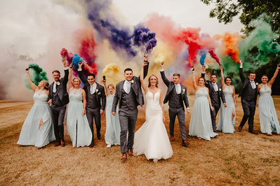 large bridal party smiling and having fun whilst walking with colourful smoke bombs