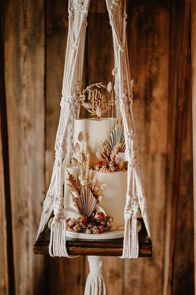 wedding cake with dried florals on a rope swing in front of a wooden wall panel in silchester barn