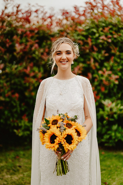 Bride, standing in a long Lacey white wedding dress, holding sunflowers, smiling
