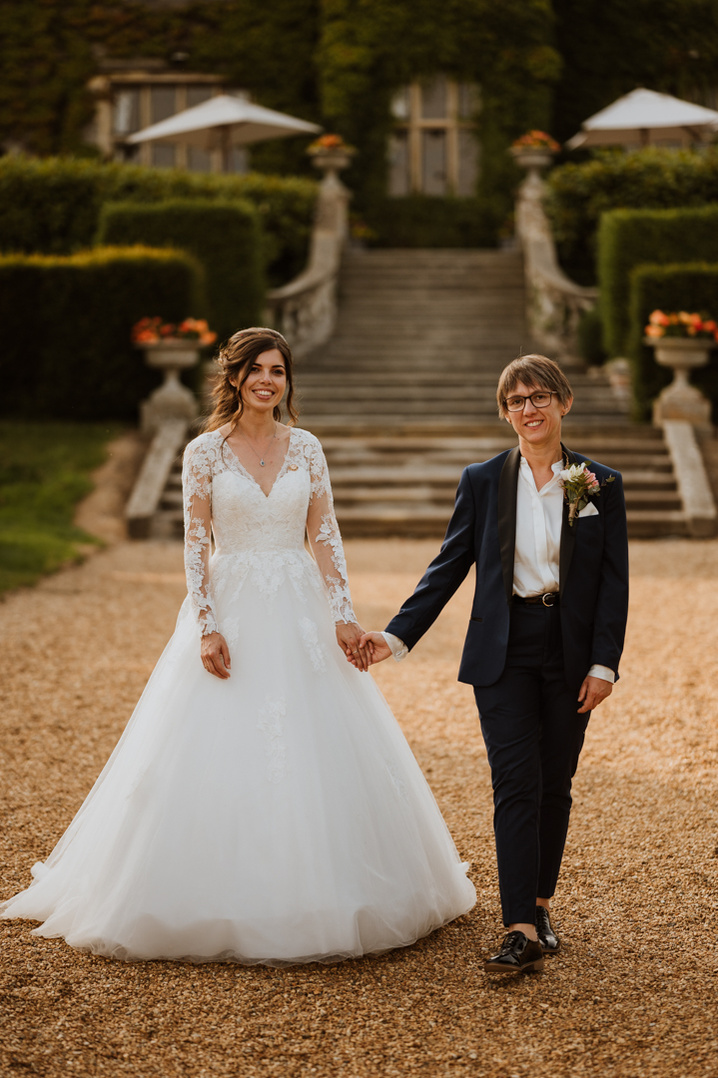 wedding couple smile to the camera whilst holding hands walking through the grounds during golden hour with the kent venue eastwell manor in the background