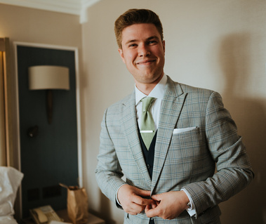 groom in a london hotel room smiling at camera in a check suit before wedding