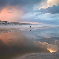 A woman looking for shells on the beach, clouds reflecting in tide pools 