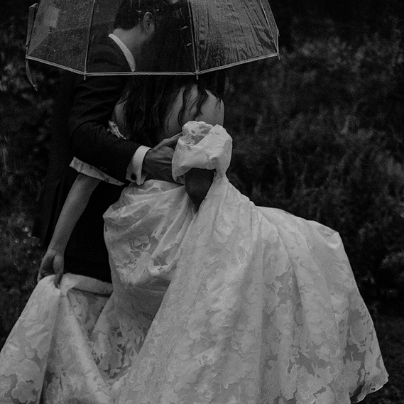 Wedding couple walking in the rain with a clear umbrella. They are holding hands and walking away from the camera. Photographed in Black and White by Hudson Valley Wedding Photographer. Location, Beacon, New York.