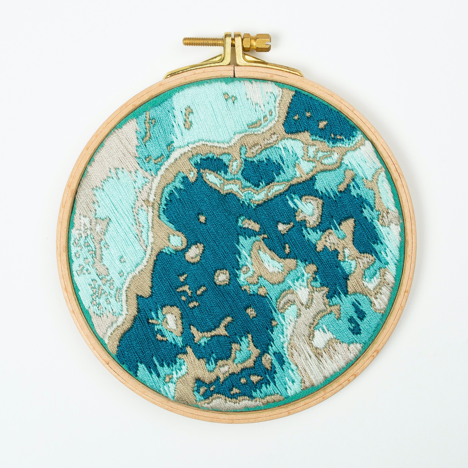 A hand embroidered art piece framed in a round, wooden embroidery hoop. The vibrant colours of the piece depict a satellite view of the ocean.