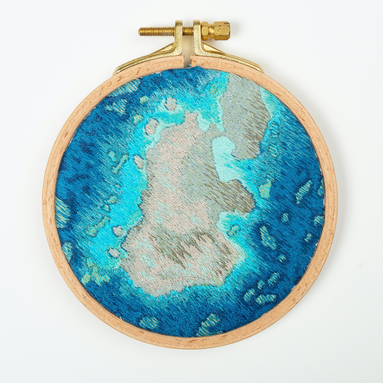 A hand embroidered art piece framed in a round, wooden embroidery hoop. The vibrant colours of the piece depict a satellite view of the ocean.