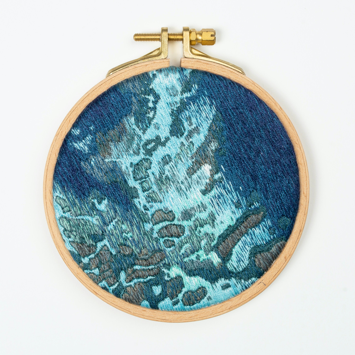 A hand embroidered art piece framed in a round, wooden embroidery hoop. The vibrant colours of the piece depict a satellite view of the ocean.