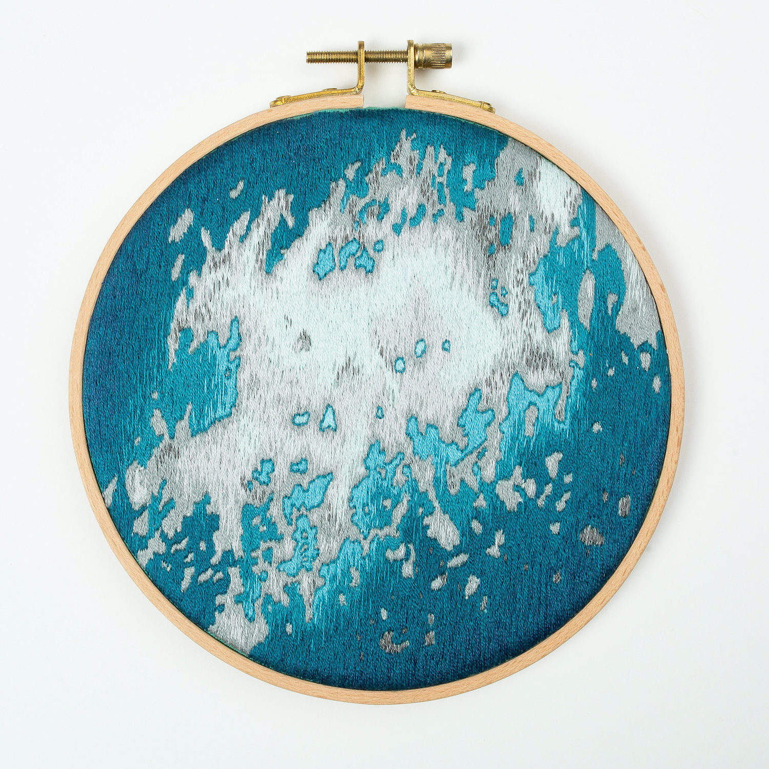 A hand embroidered art piece framed in a round, wooden embroidery hoop. The vibrant colours of the piece depict a satellite view of the ocean.