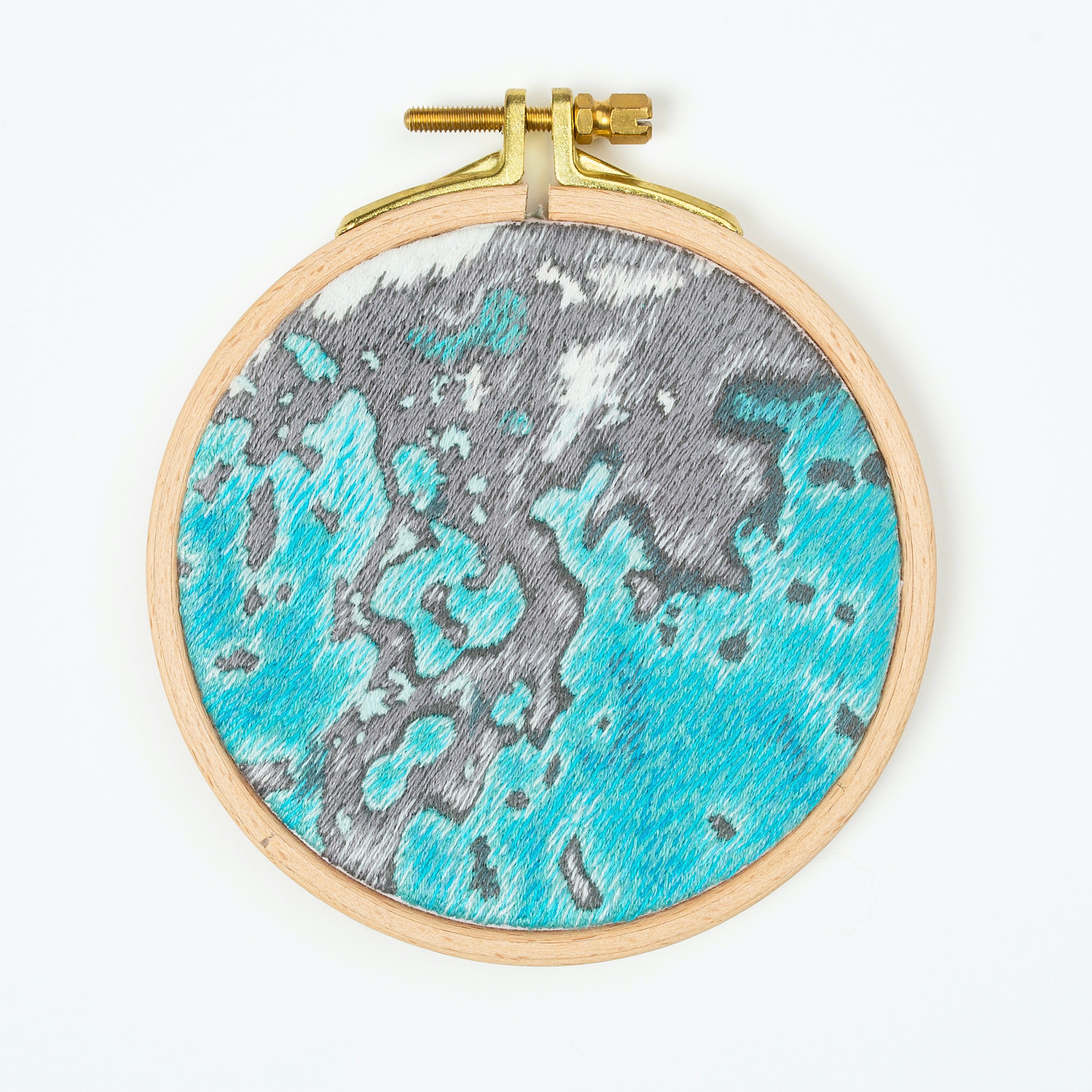 A hand embroidered art piece framed in a round, wooden embroidery hoop. The vibrant colours of the piece depict a satellite view of the ocean.