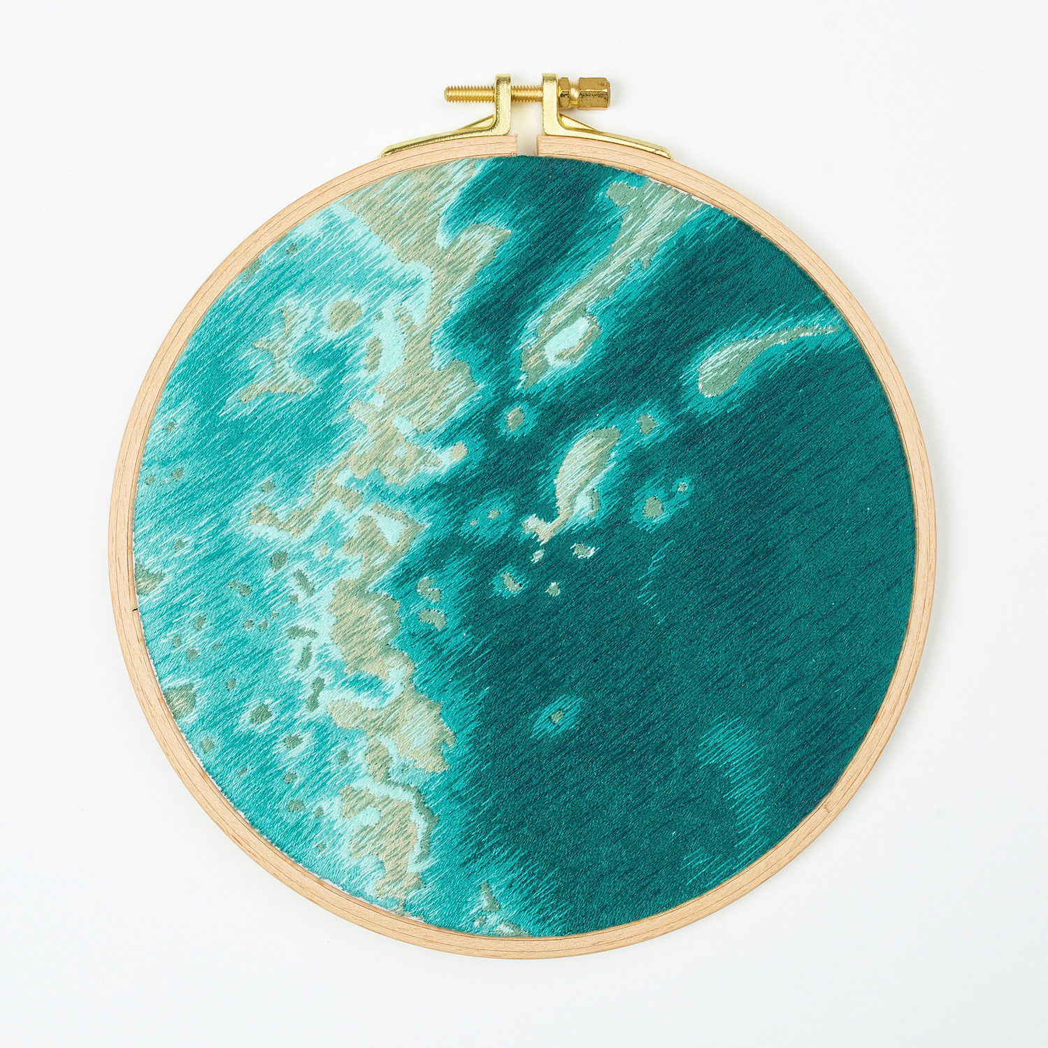 A hand embroidered art piece framed in a round, wooden embroidery hoop. The vibrant colours of the piece depict a satellite view of the ocean.