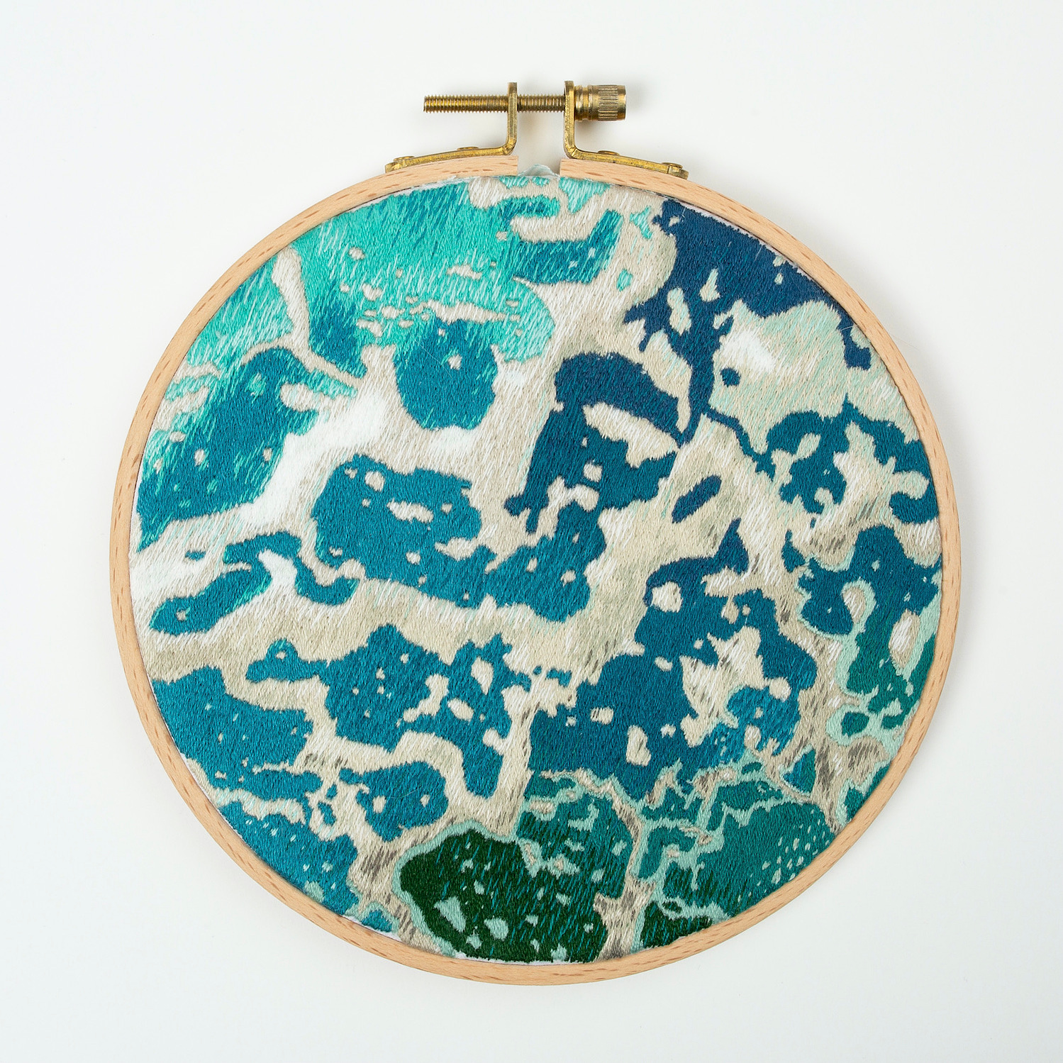 A hand embroidered art piece framed in a round, wooden embroidery hoop. The vibrant colours of the piece depict a satellite view of the ocean.