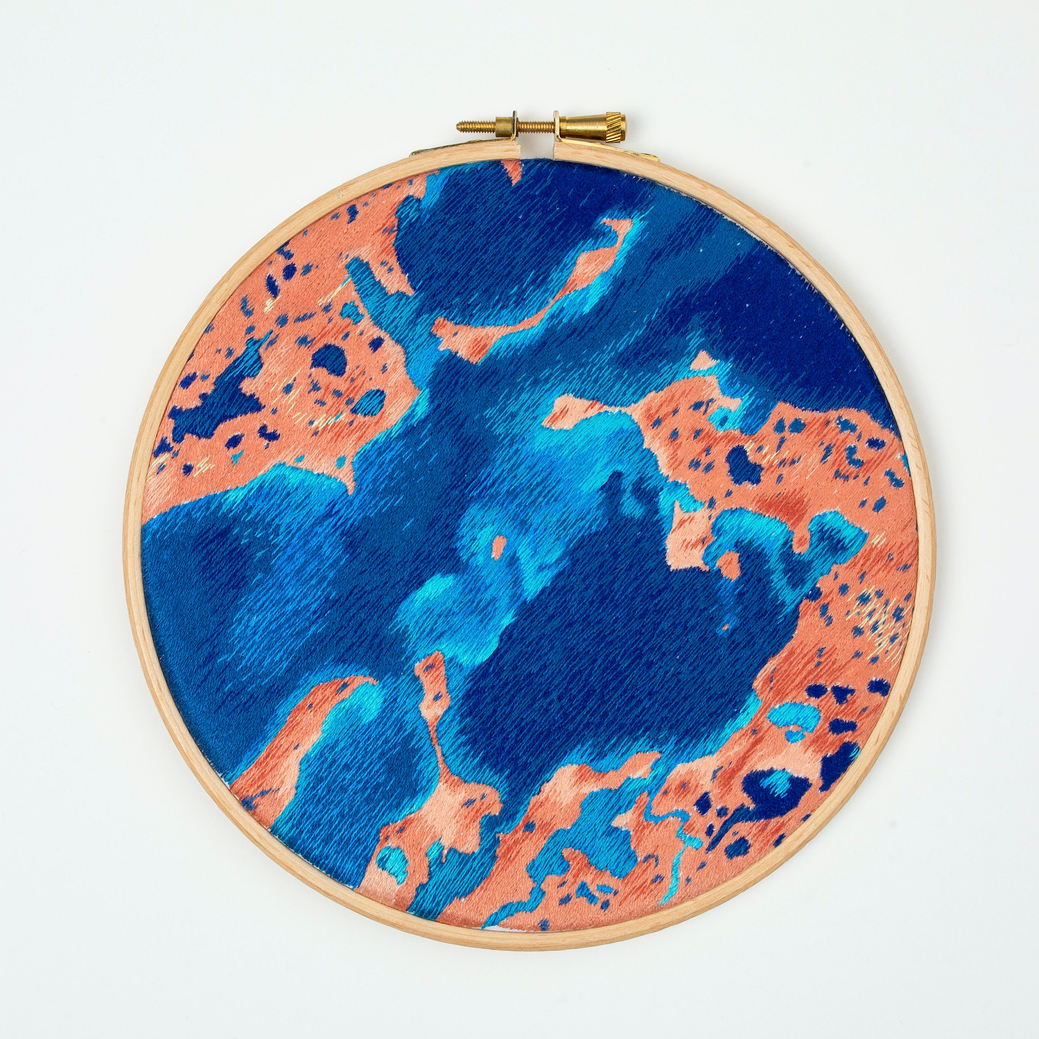 A hand embroidered art piece framed in a round, wooden embroidery hoop. The vibrant colours of the piece depict a satellite view of the ocean.