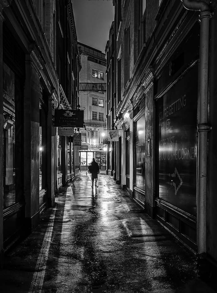 A black and white photo of a figure walking at night in the City by Mark Burley