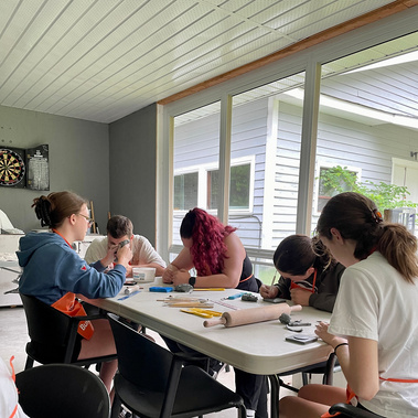 A photo of a group of young adults around a craft table on the porch of Lake Joe's Rec Hall. They are all engaged with making their own tiles, working with rolling pins, ceramic tools and their hands. They wear aprons, and none are looking at the camera.