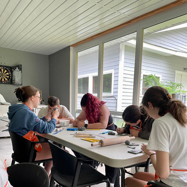 A photo of a group of young adults around a craft table on the porch of Lake Joe's Rec Hall. They are all engaged with making their own tiles, working with rolling pins, ceramic tools and their hands. They wear aprons, and none are looking at the camera.