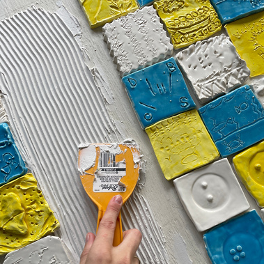 A photo of the process of making the mural: a table with the mostly-completed mural, with a hand holding a scraper tool to apply the putty used to mount the tiles to the backing board.