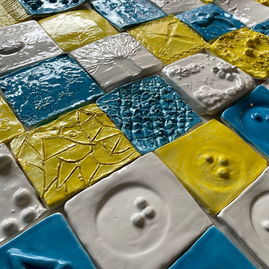 A similar photo to the previous one, but this one is closer to the mural, showing off the relief of the texture. Some braille characters are at the bottom, some tiles are abstract textures, and one is of a tree. They are glazed in blue, yellow and white.