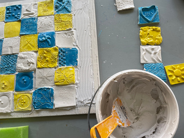 A photo of the process of making the mural: a table with the mostly-completed mural, with the putty in a bucket that was used to mount the tiles to the backing board.