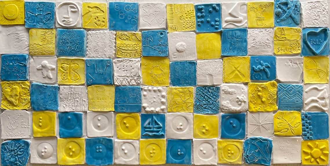 A mosaic mural made up of squares, each is different. They are made of clay and are decorated with various tactile artworks. The tiles are different colours: yellow, blue and white, in a repeating pattern.